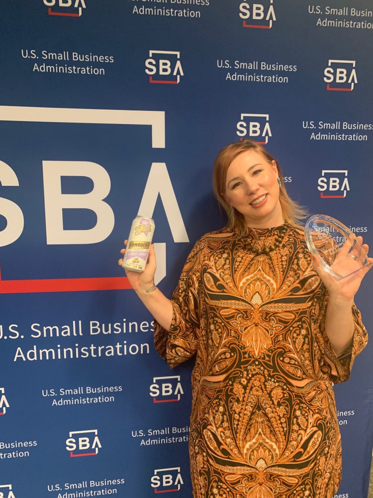 woman poses with trophy and a can of kombucha in front of SBA banner