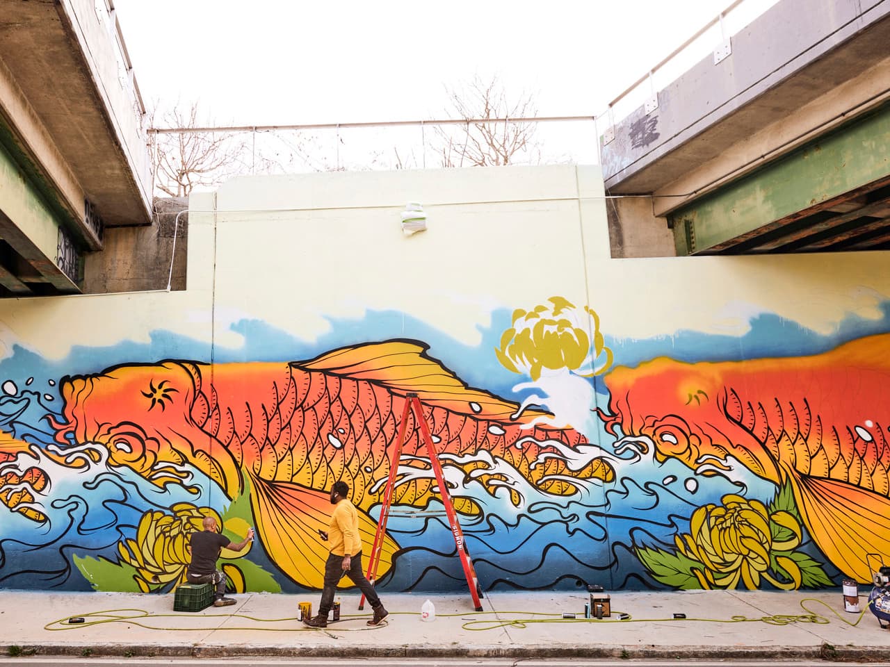 A man sits with his back toward the camera, painting a large colorful mural of fish and water. Another man walks by the artist at work.