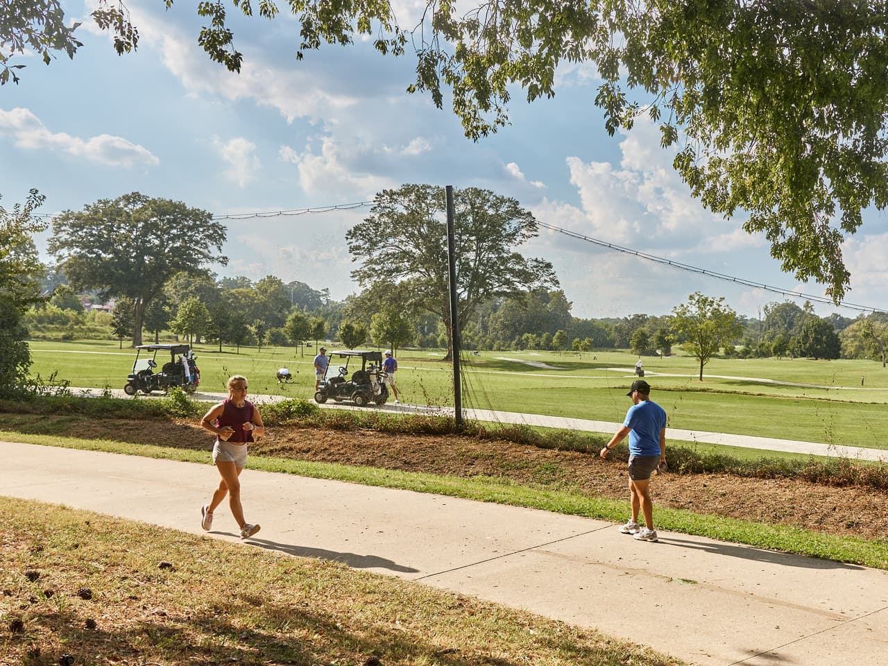 People jogging and walking on a path beside a golf course with golf carts and trees under a blue sky.