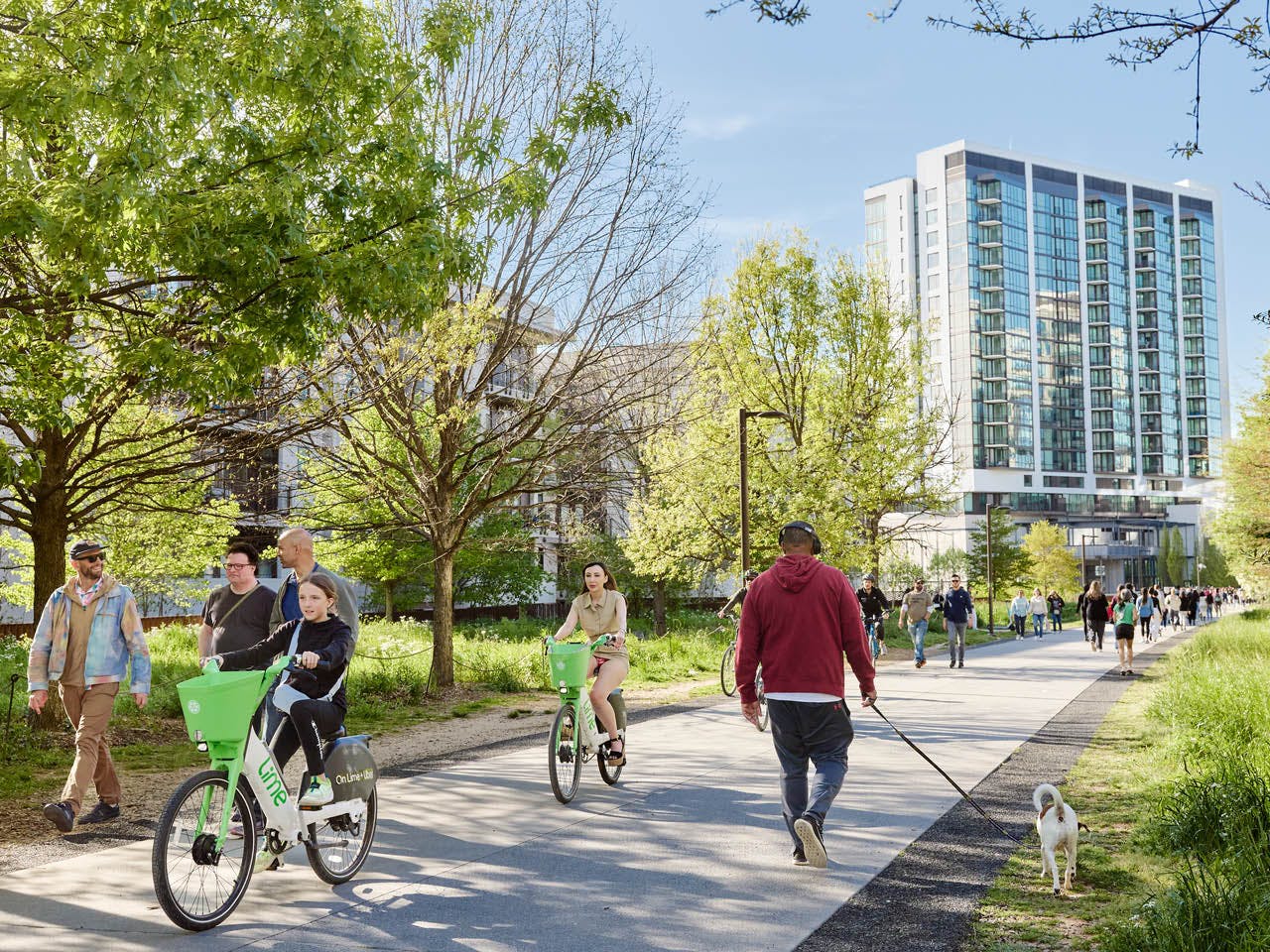 People ride bikes and walk dogs on the Eastside Trail with trees and buildings in the background. (Photo Credit: Erin Sintos)