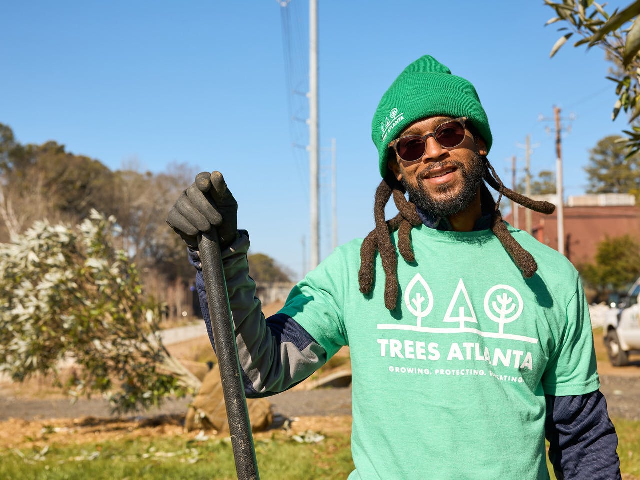 A man stands, wearing a Trees Atlanta shirt and holding a shovel.