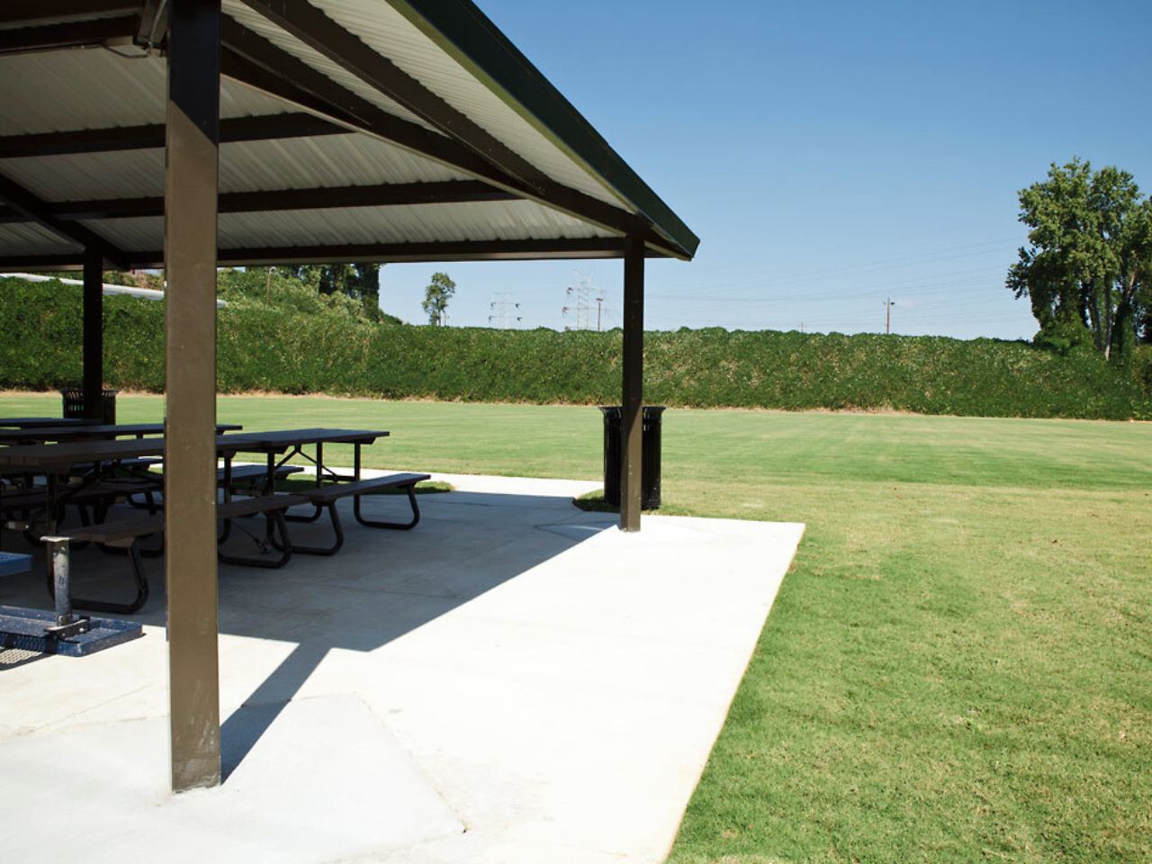 A covered pavilion sits in a green grass field with picnic tables underneath.