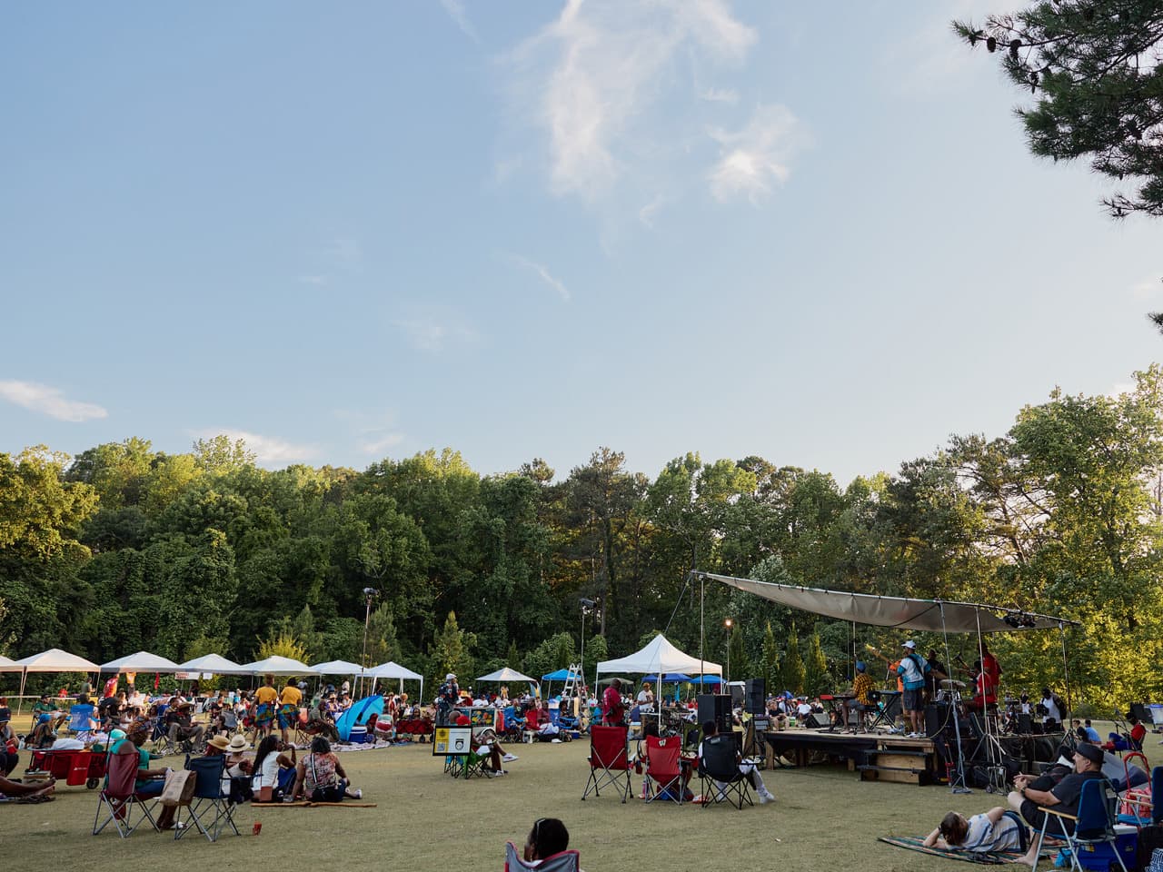 Crowds of people sit in a park in chairs and on blankets watching a performance as the sun sets.