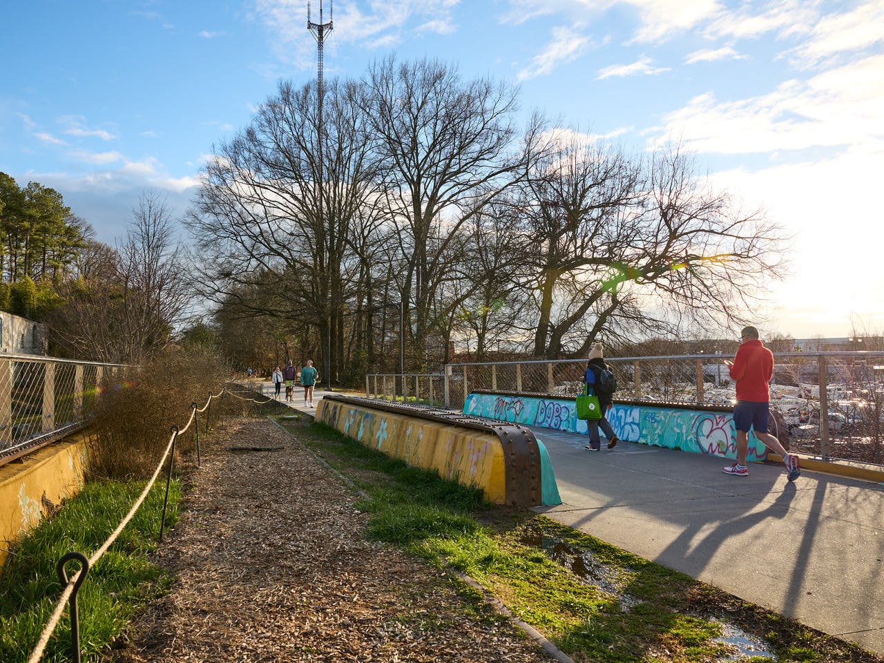 A man jogs across a bridge. In front of him, a woman walks with groceries. Other people walk in the background.