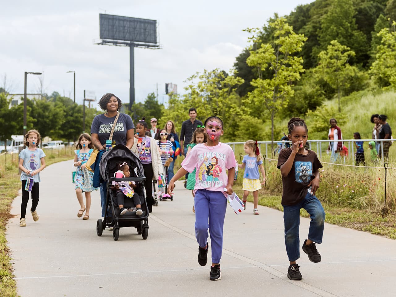 A group of children parade down the Beltline, with one child in a stroller and others holding small items, surrounded by green trees.
