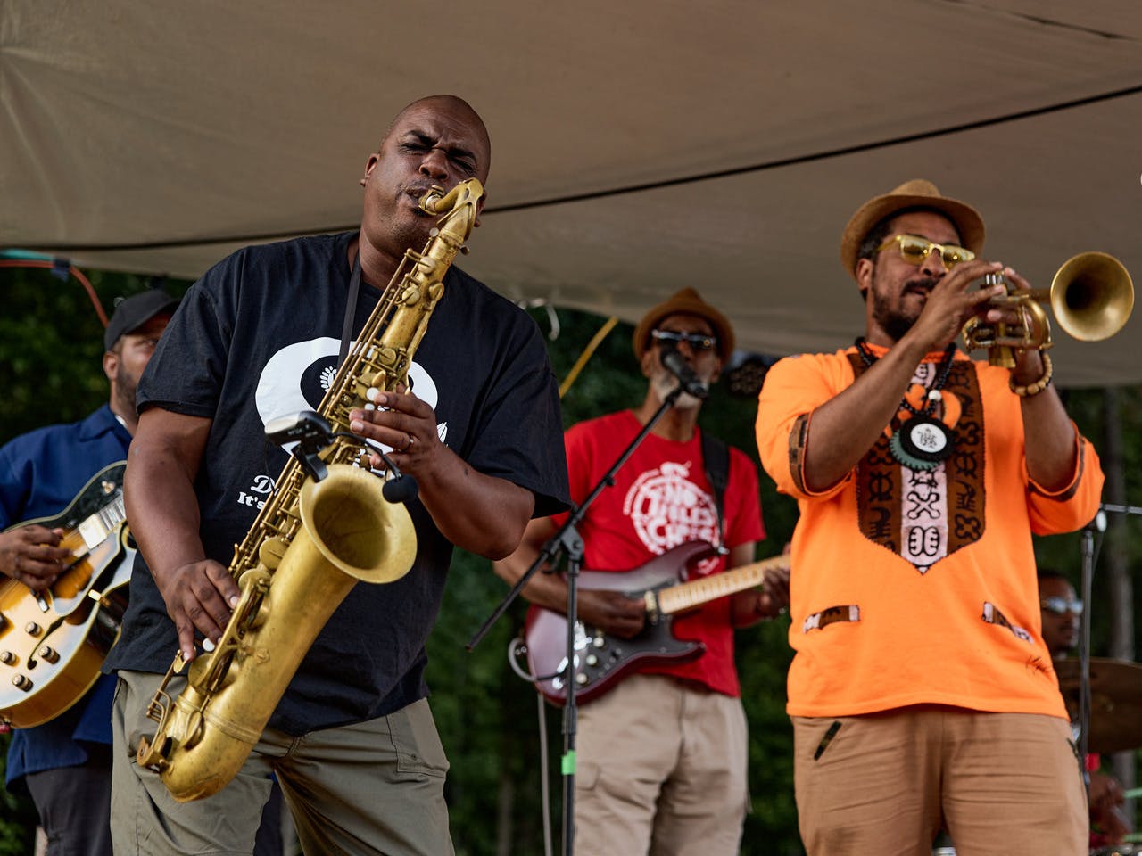 A band performs. In the front, one man plays a tenor saxophone next to a man playing the trumpet. Behind them, two men have guitars.