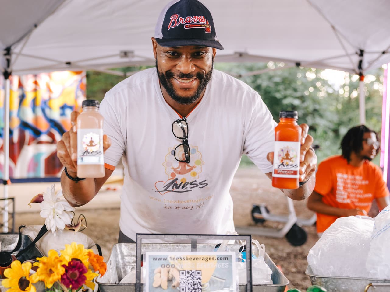 Man under a tent smiling, holding bottles of juice labeled "1ness." Flowers and a QR code are on the table. Another person sits in the background.