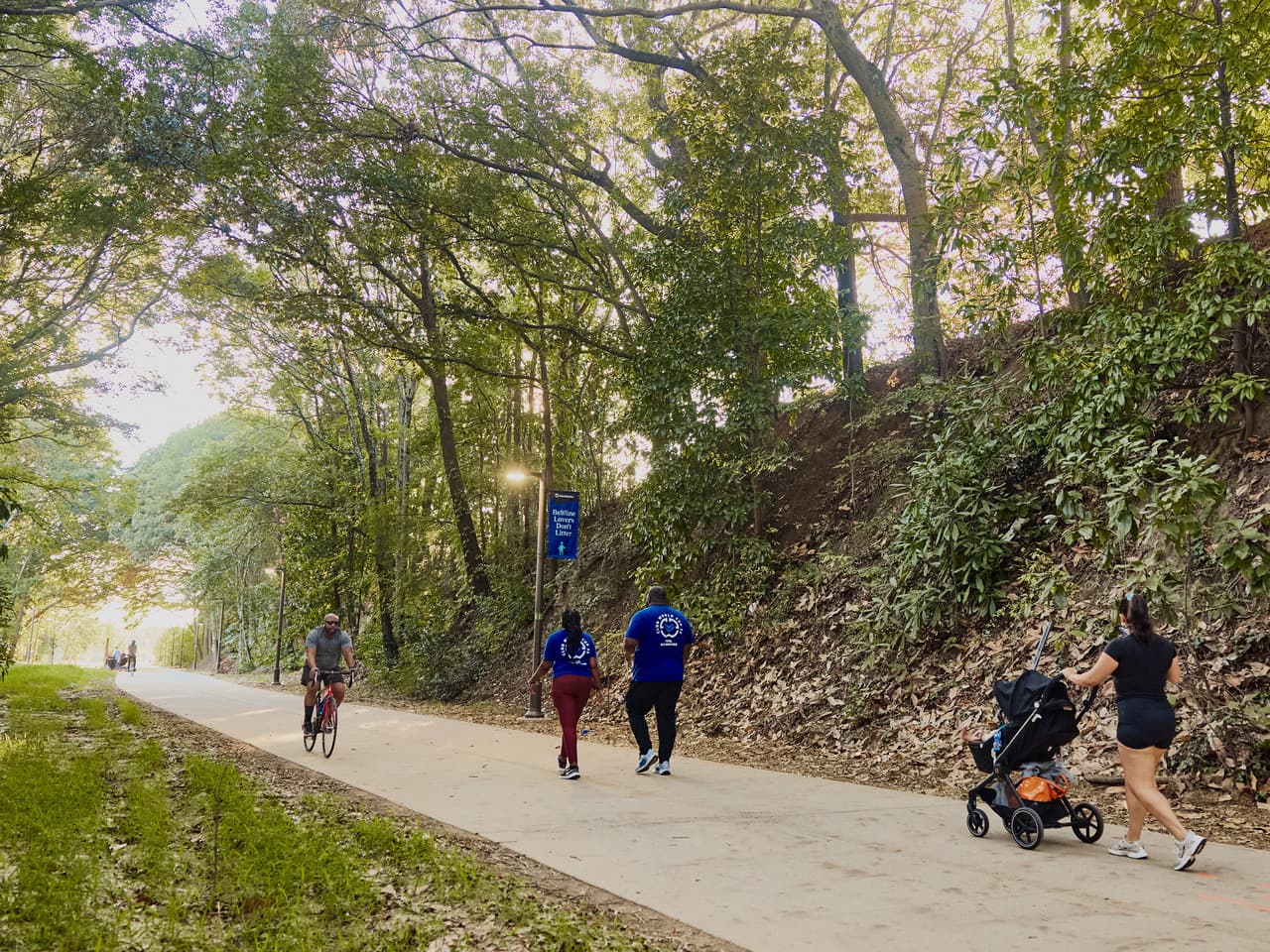 People walking, jogging, and biking along a tree-lined path; woman pushes a stroller.