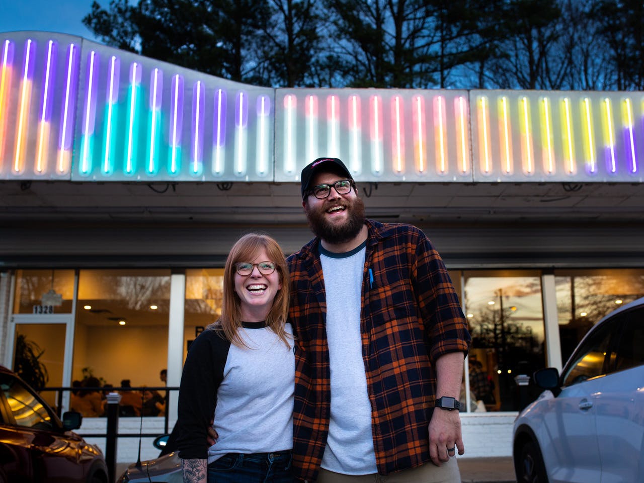 A woman and a man stand together, smiling, in front of a building with bright neon lights atop it.