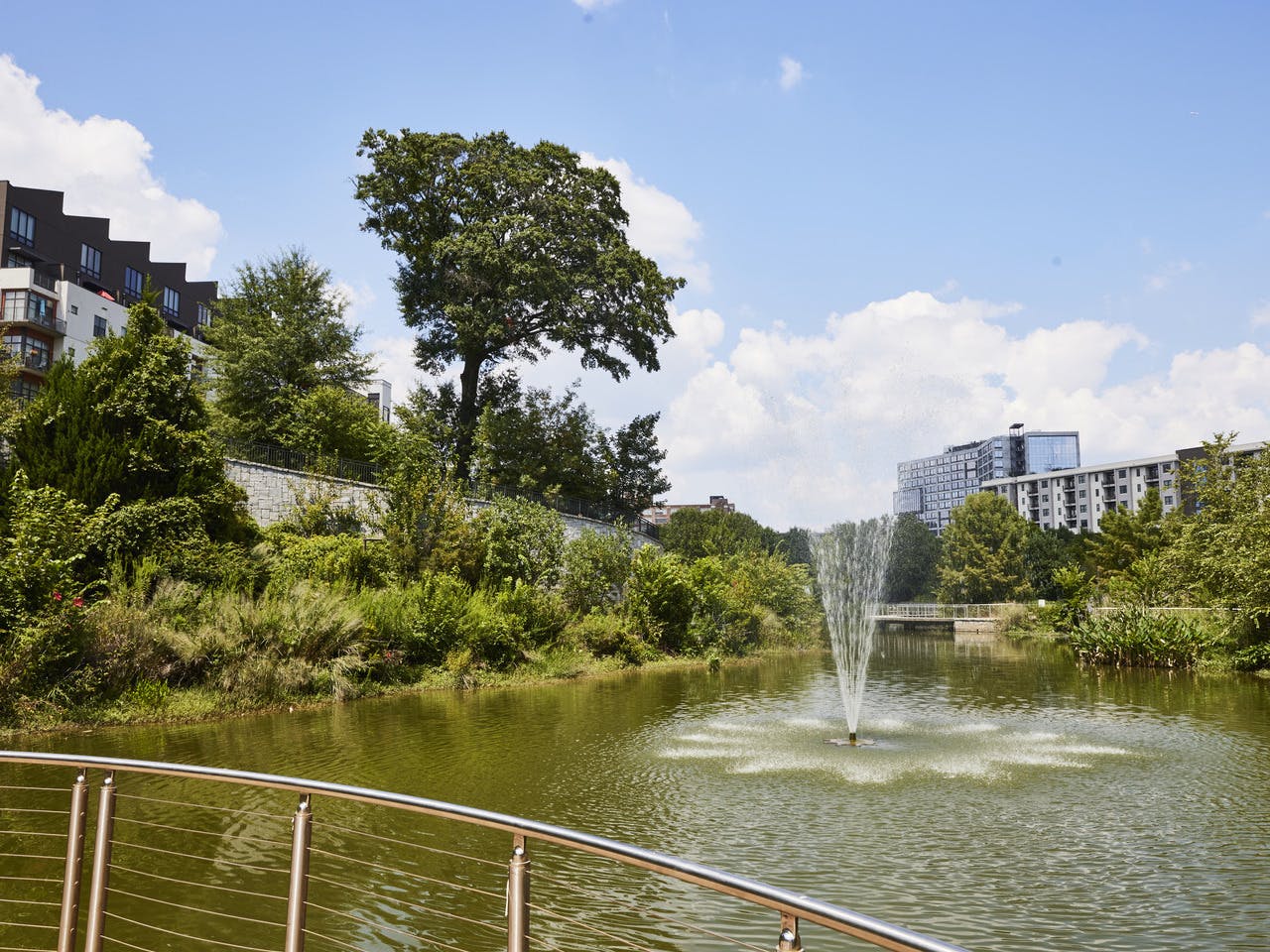 A water feature is located in a large pond with greenery and buildings around it.