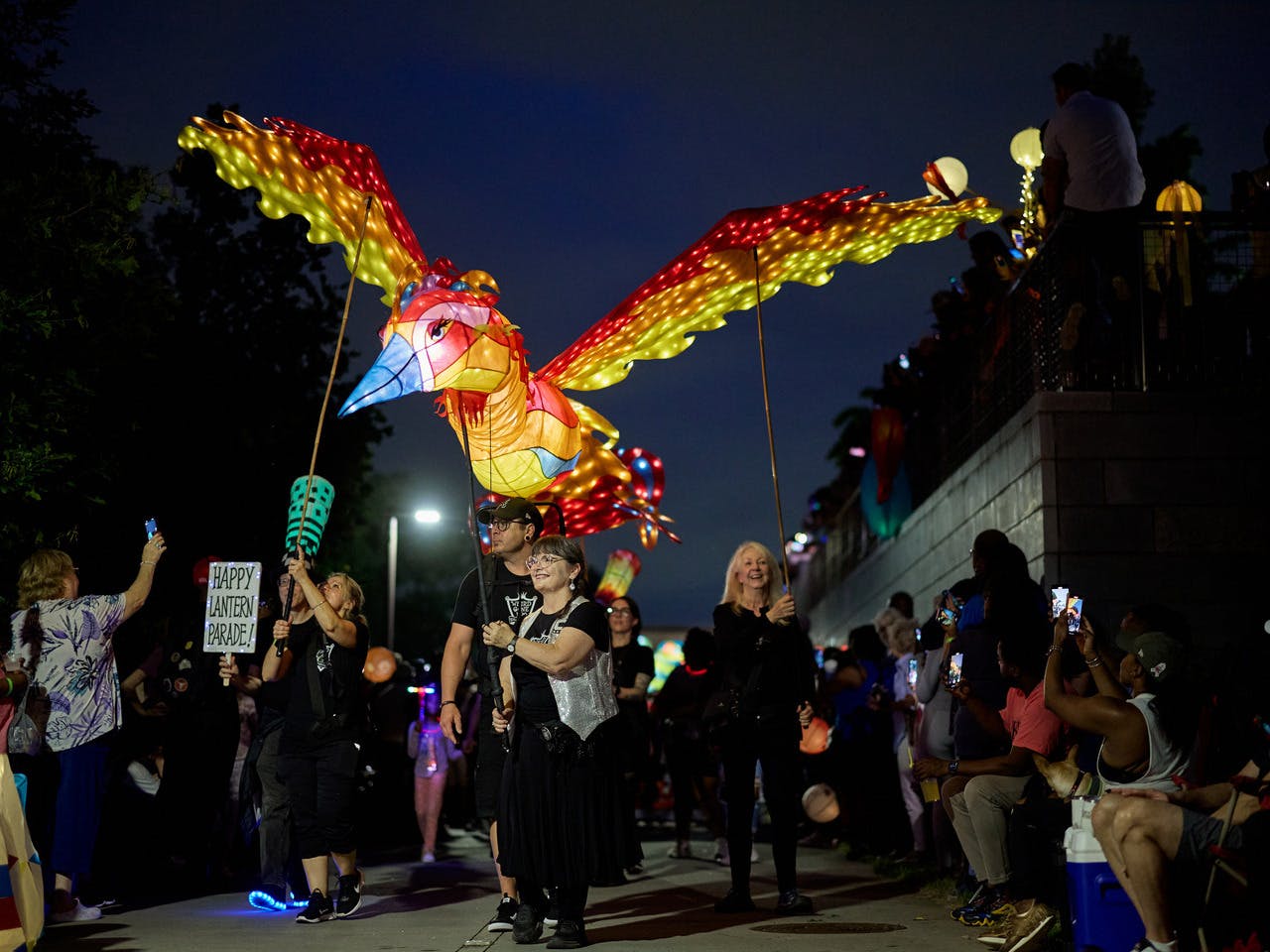 A woman walks, holding a giant, vibrantly colored, and illuminated phoenix lantern puppet. A crowd stands on either side of her.