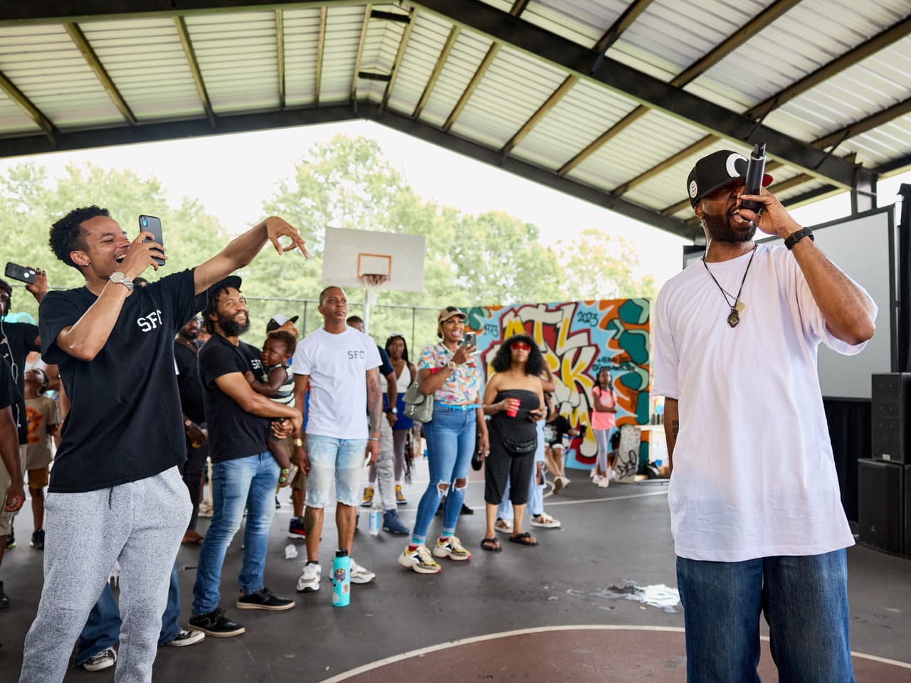 A man stands to the right, rapping into a microphone, while a crowd lines up to his left cheering him on and filming.