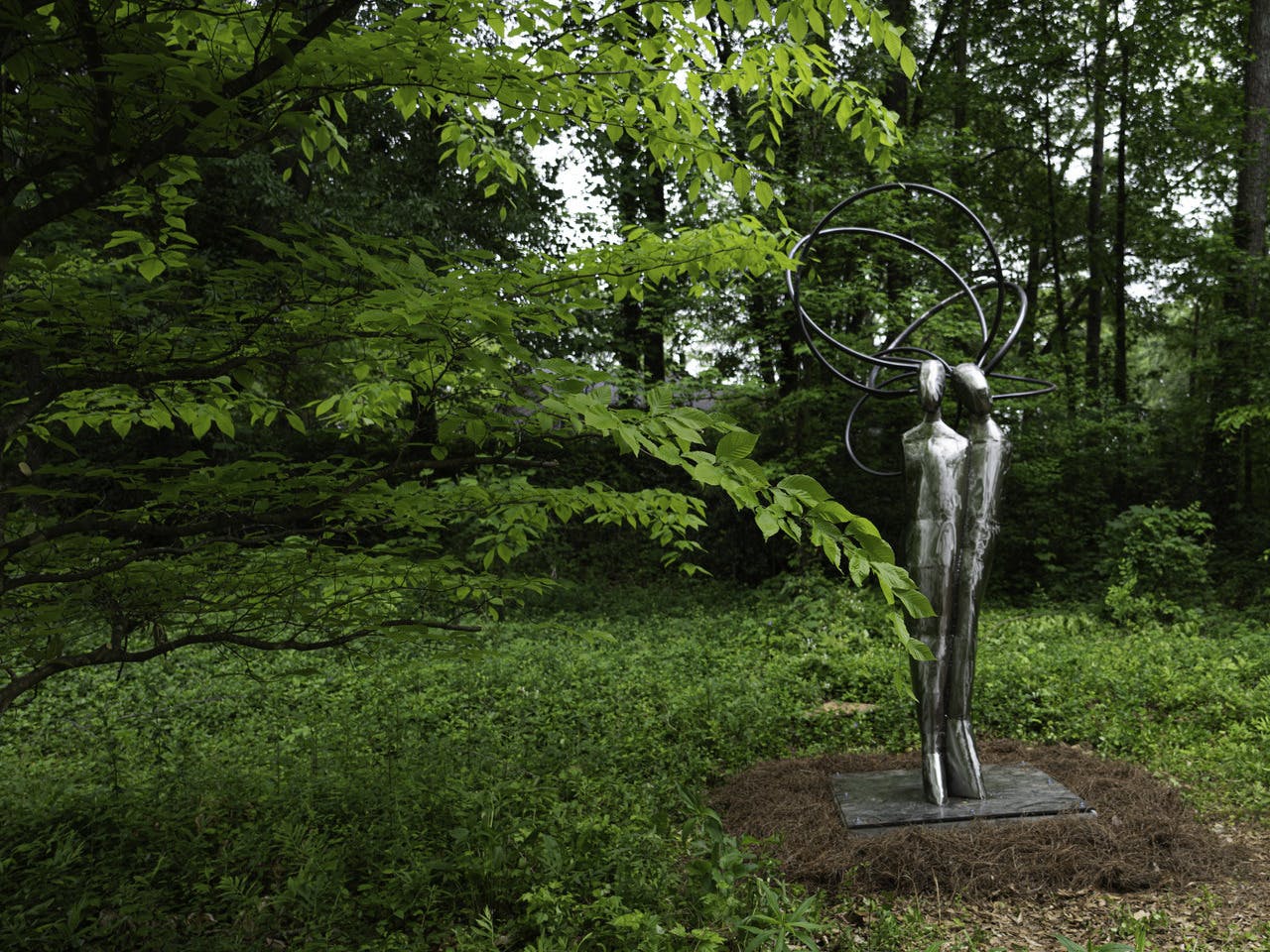 A silver statue of two twin women sits in a greenspace surrounded by trees.