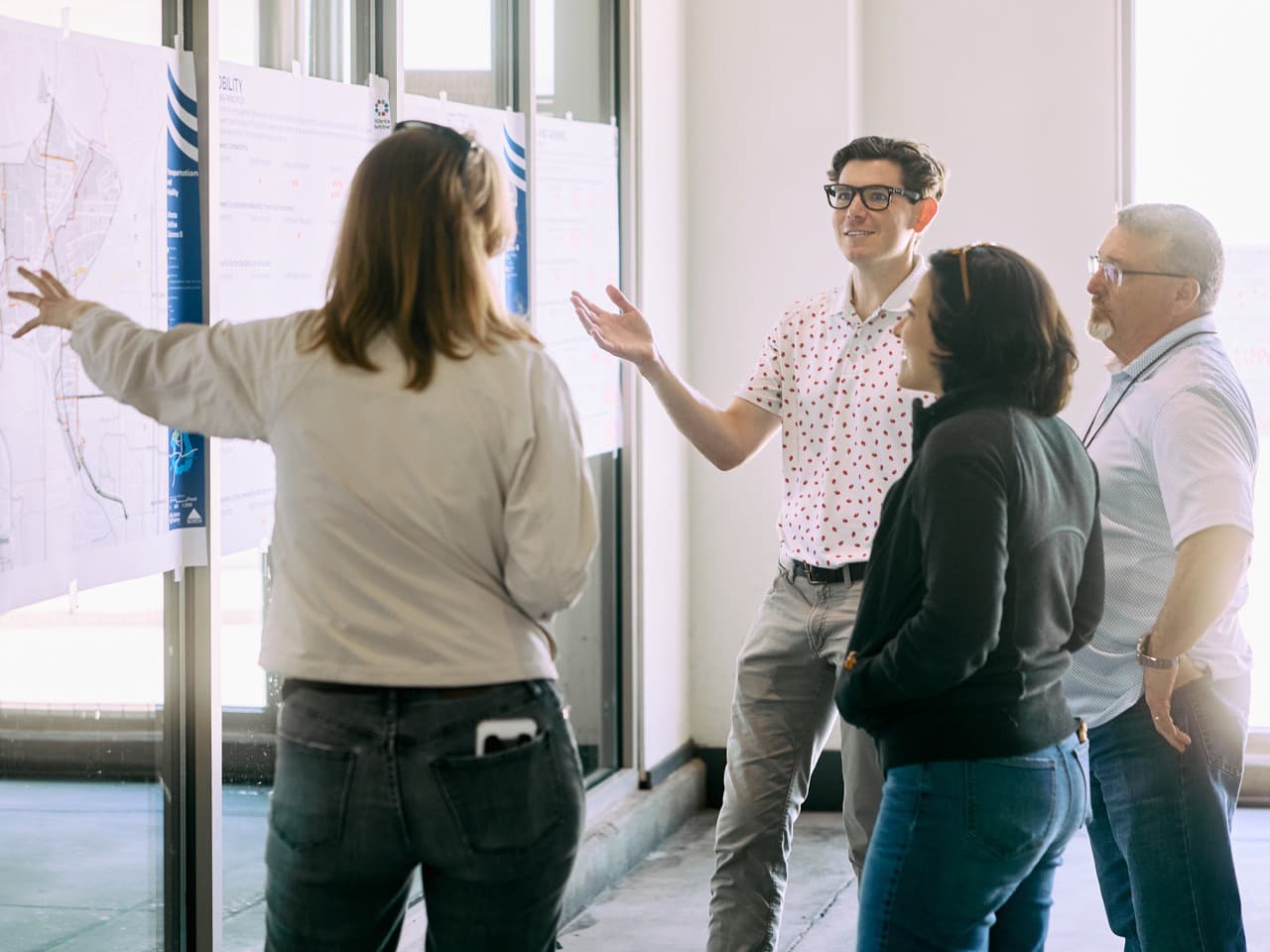 Four people engaged in discussion, reviewing maps and charts on a wall in a bright room. One person gestures towards the display.