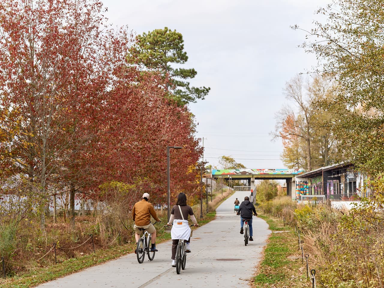 A group of people bike down a paved path, surrounded by trees adorned with autumn-colored leaves.