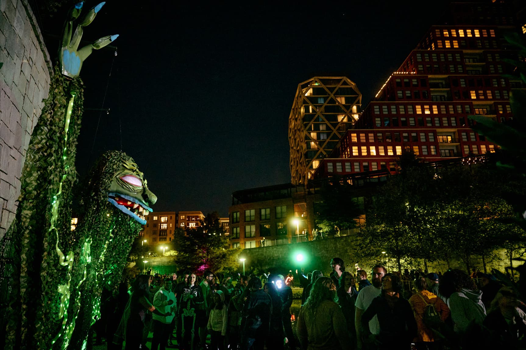Night event with a large green puppet, illuminated buildings, and a crowd of people enjoying the lively atmosphere.