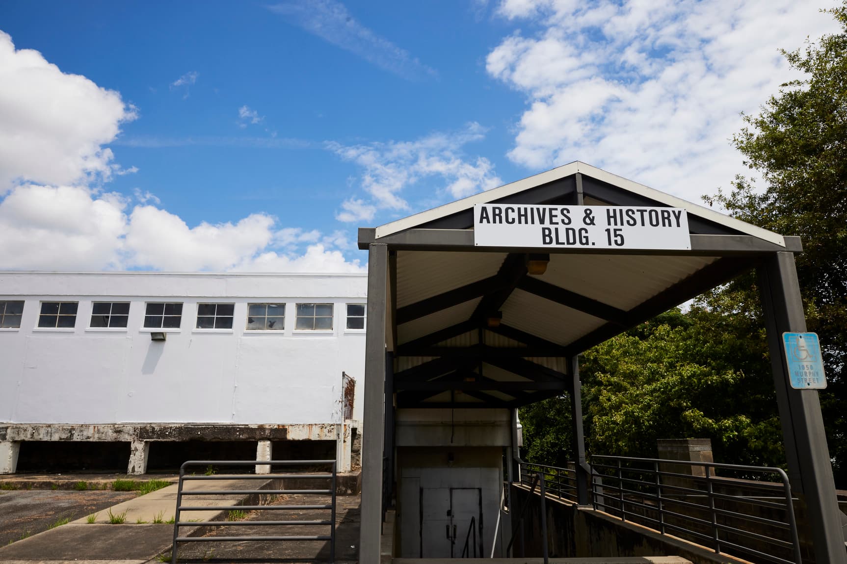 Entrance to "Archives & History Bldg. 15" with a covered walkway, surrounded by a white building and trees under a partly cloudy sky.