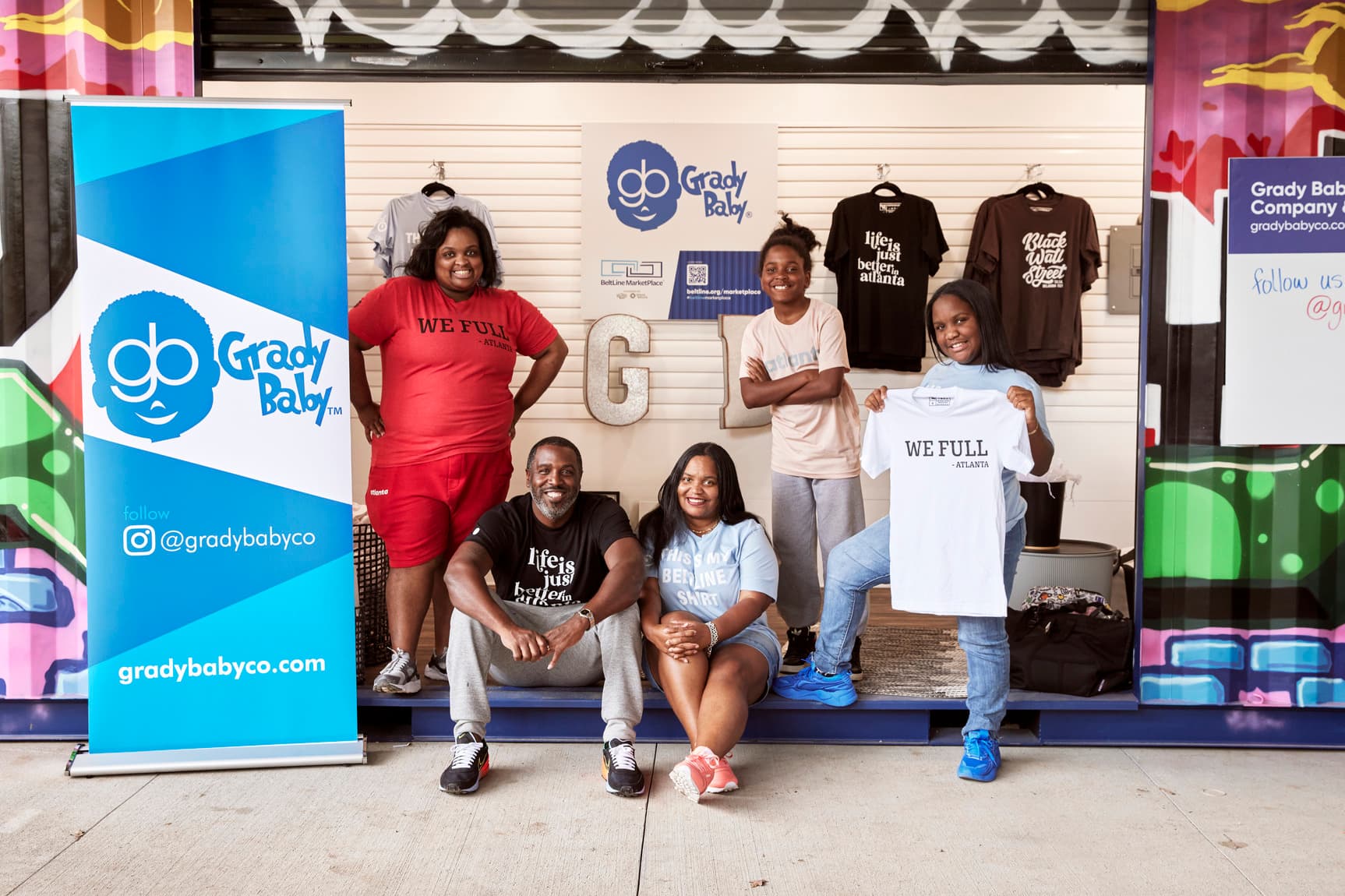 A family stands in front of a container repurposed as a store smiling and holding shirts for sale.