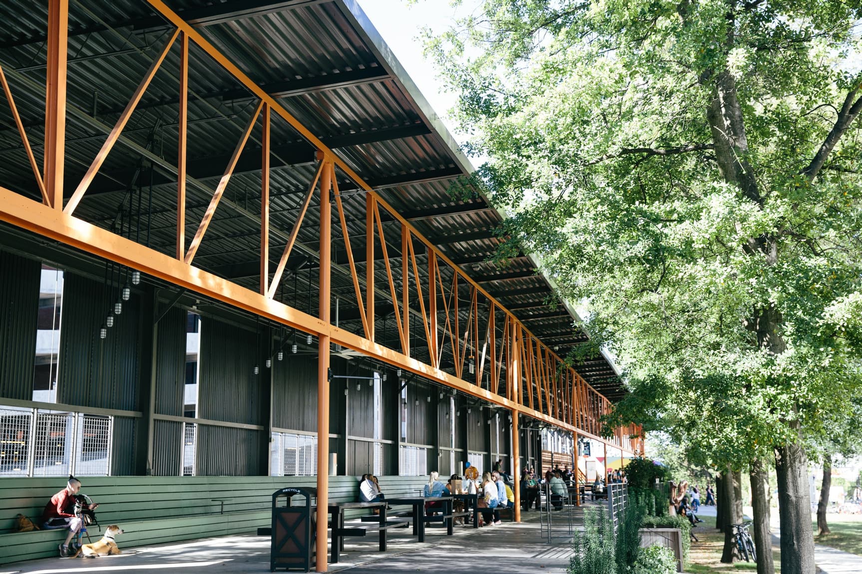 A photo of an open-air outdoor structure made from metal siding, orange pipes, and more. People sit along green benches and trees grow to the side.