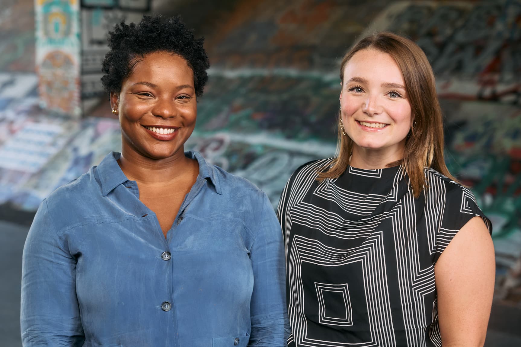 Two people stand, smiling at the camera, with a graffiti mural behind them.