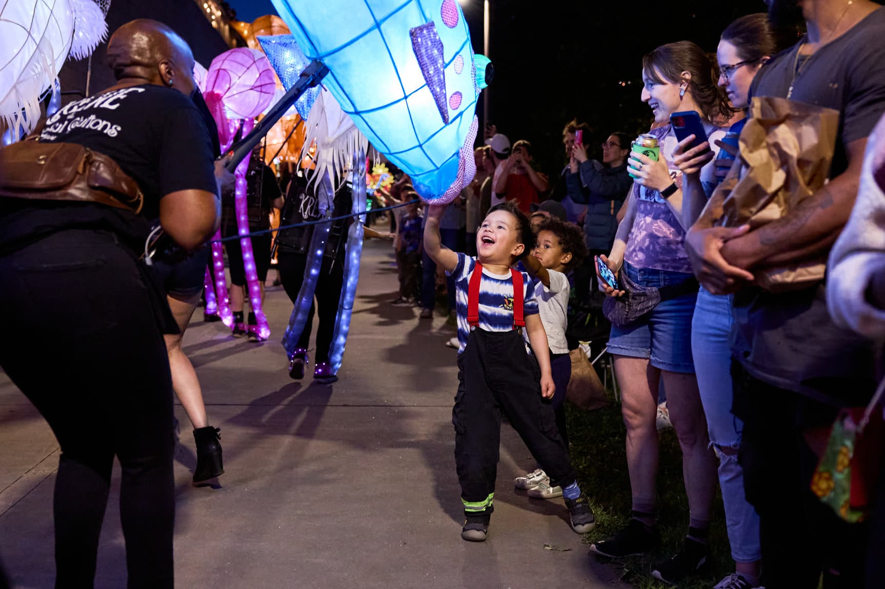 A child excitedly reaches for a glowing lantern held by a performer during a lively parade, with a crowd watching and smiling.