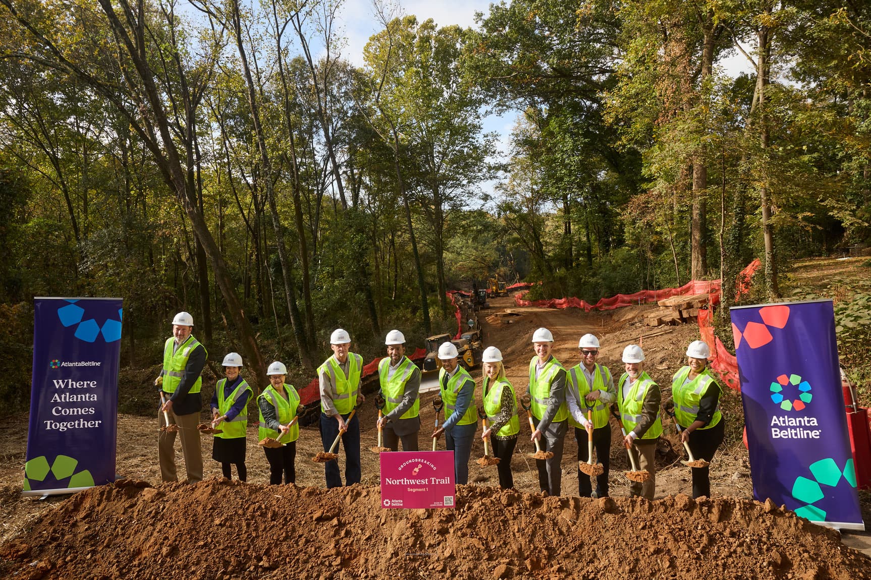 A group of people holding shovels stand behind a pile of dirt with a construction site in the back.