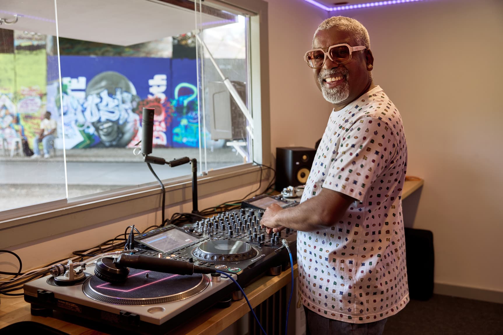 Smiling older man DJing at turntables and mixer in a studio, wearing glasses, with colorful graffiti mural visible through the window.