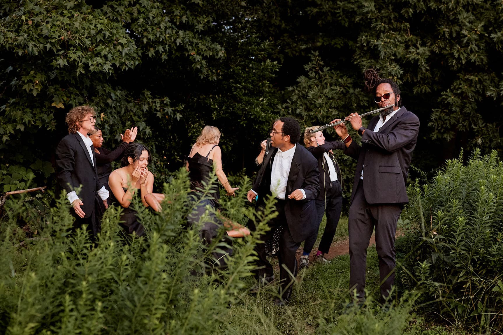 A group of people dressed in formal black attire gather in a lush, green outdoor setting, with one person playing a flute.