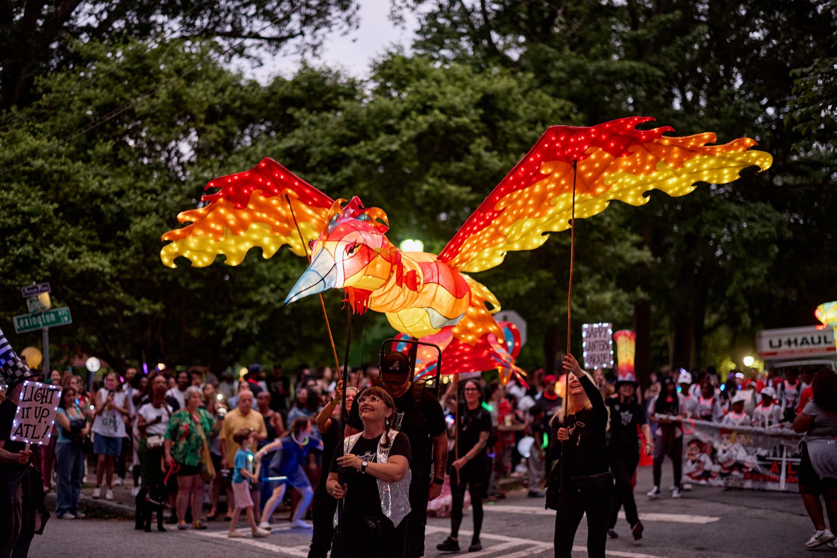 A parade with people holding a large illuminated phoenix-shaped lantern, surrounded by a crowd and trees in the background.