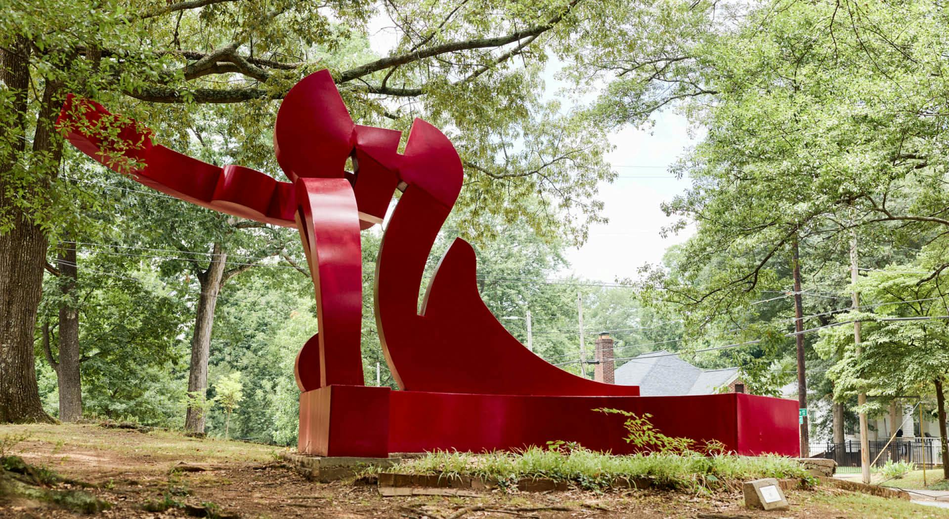 A large red abstract sculpture sits amongst the tall trees in Perkerson Park. (Photo Credit: Erin Sintos)