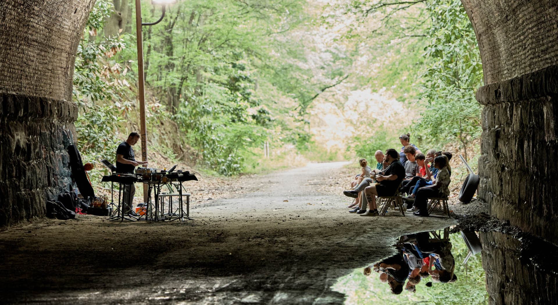 A seated audience watches a musician perform in a stone tunnel reflecting in a water puddle. (Photo Credit: Erin Sintos)