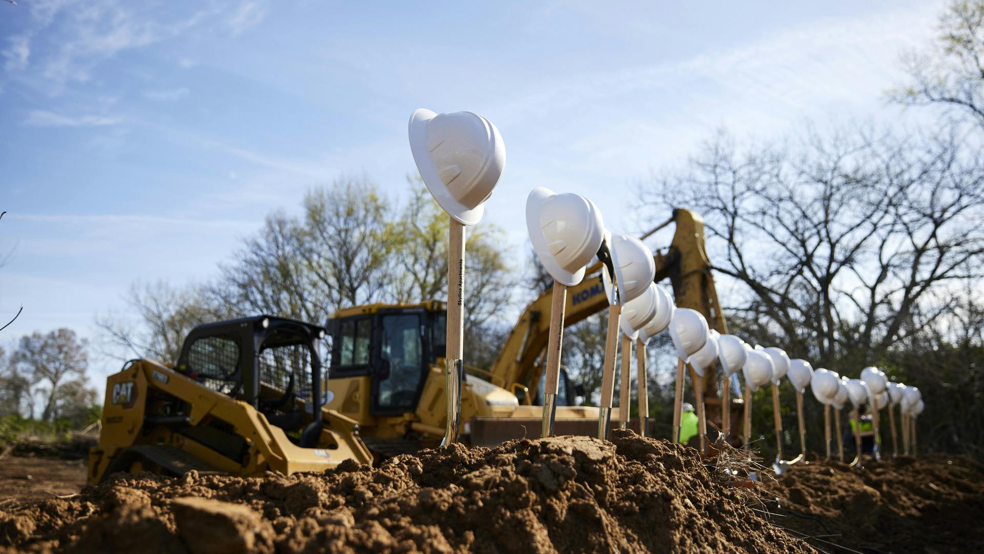 Construction equipment sits behind a line of shovels, standing in dirt, with construction hard hats balanced on top of them.