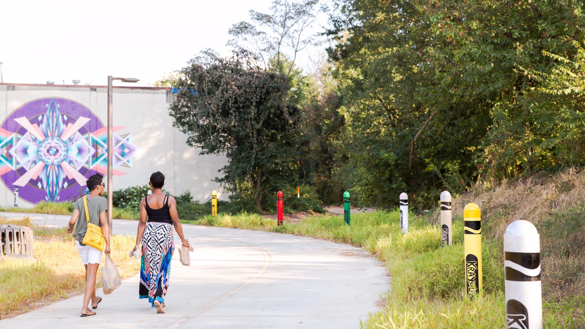 Two people walk along a paved trail that's lined with crayon statues on one side. In the distance, there is an unpaved path where the Westside Trail - Segment 6 will go.