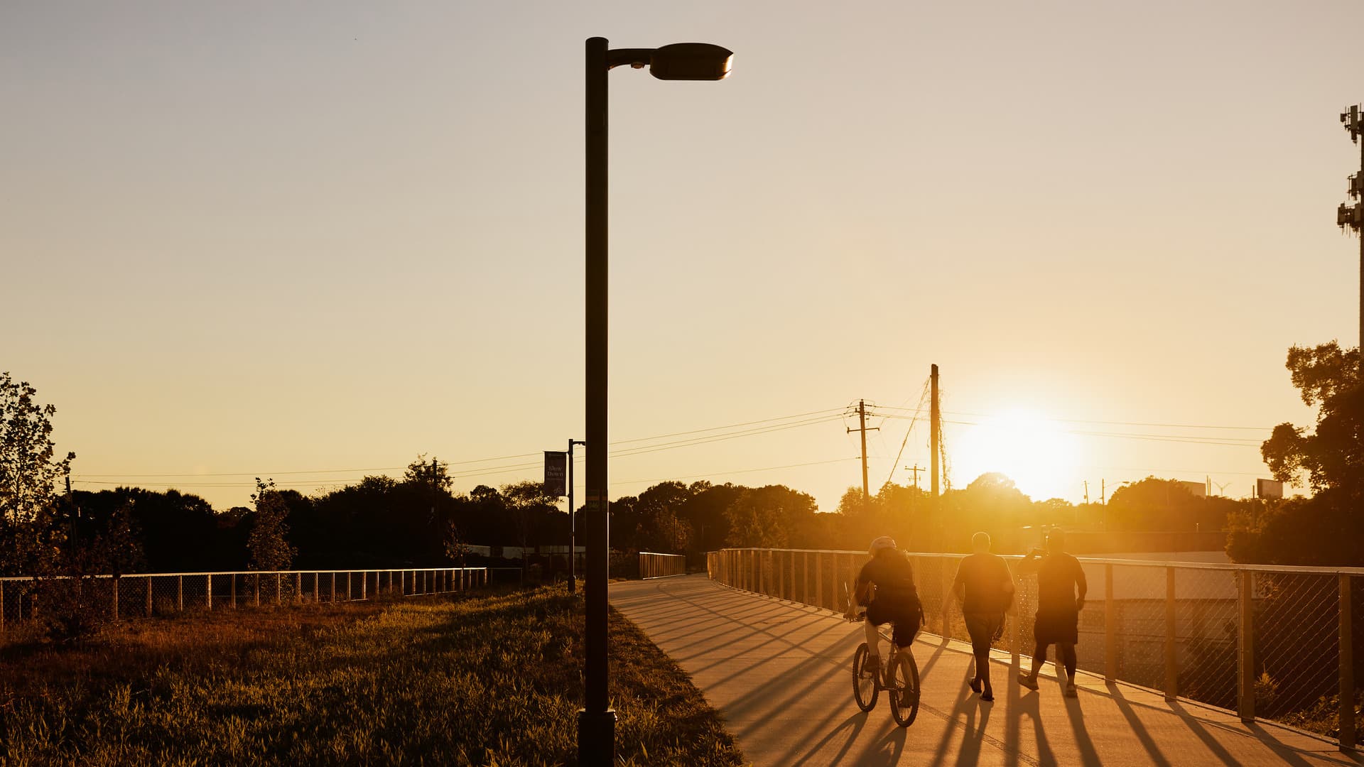 People walking and biking on a paved path at sunset, with shadows stretching across the ground and a lamppost against the sky.