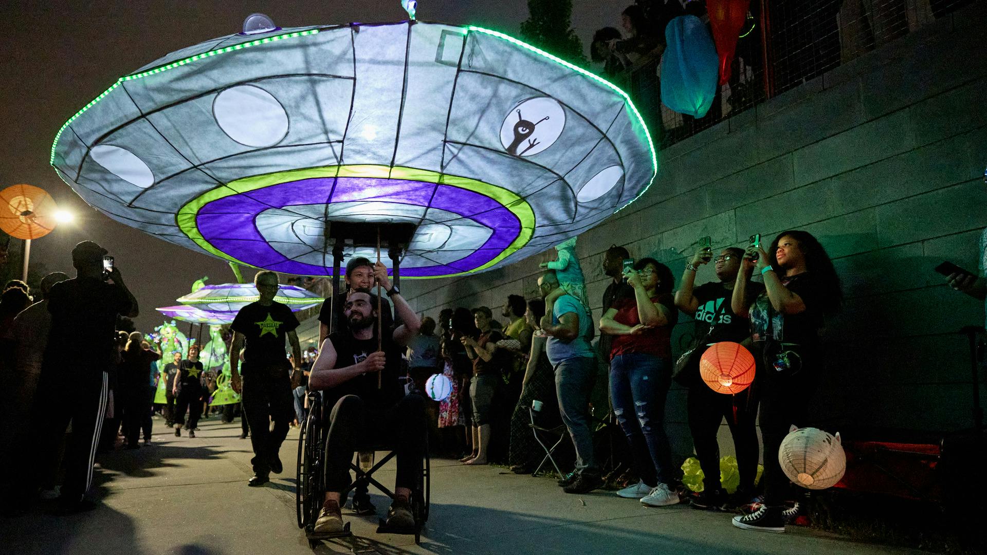 A man in a wheelchair holds a large illuminated puppet of an alien spaceship.