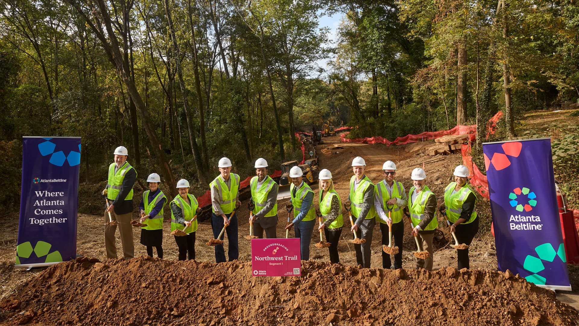 A group of people holding shovels stand behind a pile of dirt with a construction site in the back.