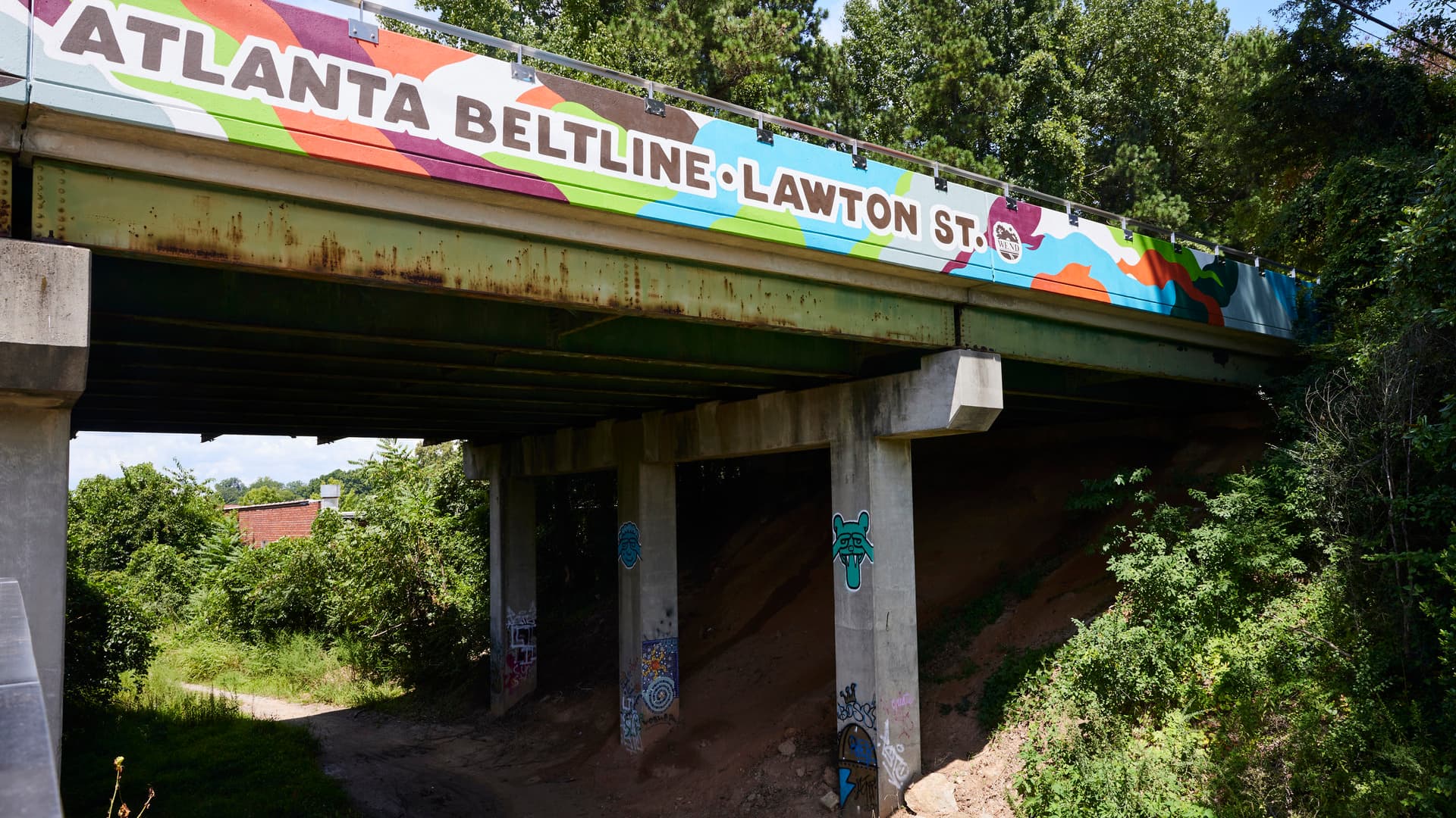 A bridge goes over an unpaved path. On the side of the bridge is a colorful mural that reads, "Atlanta Beltline. Lawton St."