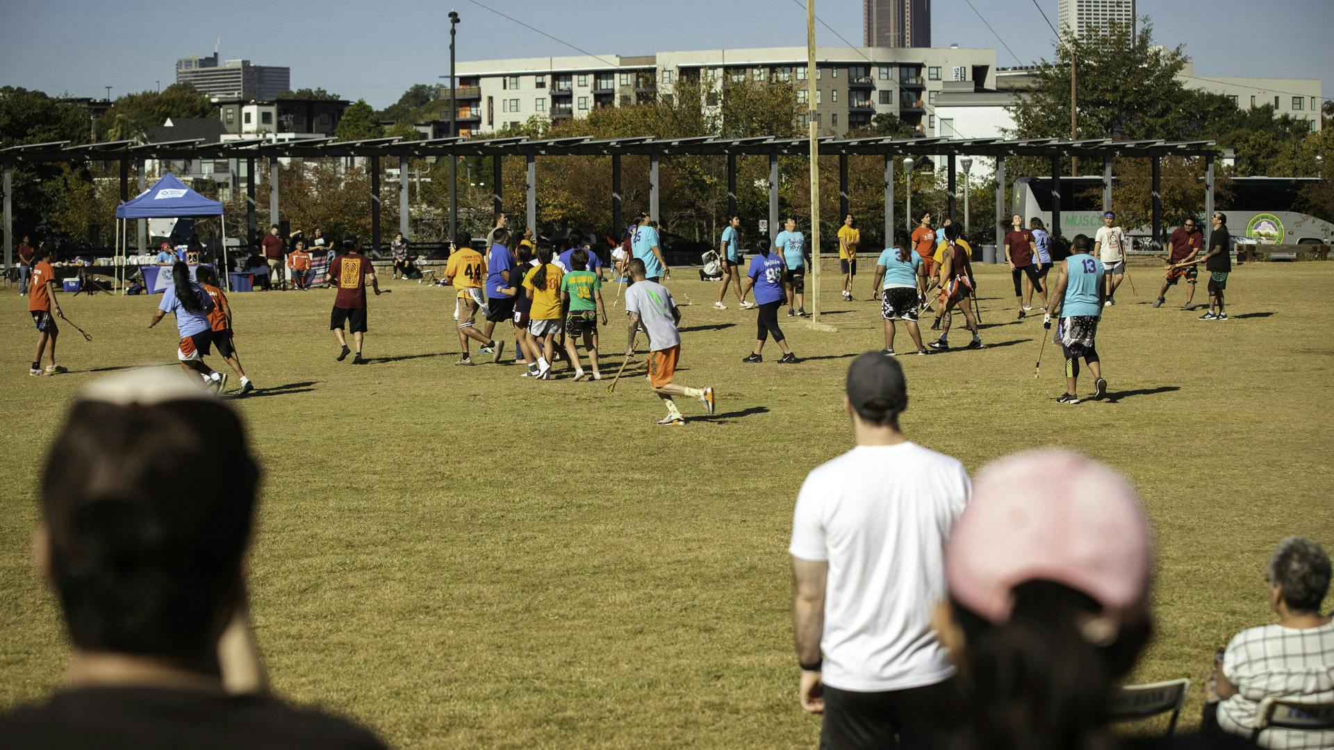 A group of people play Stickball on a field while spectators watch on.