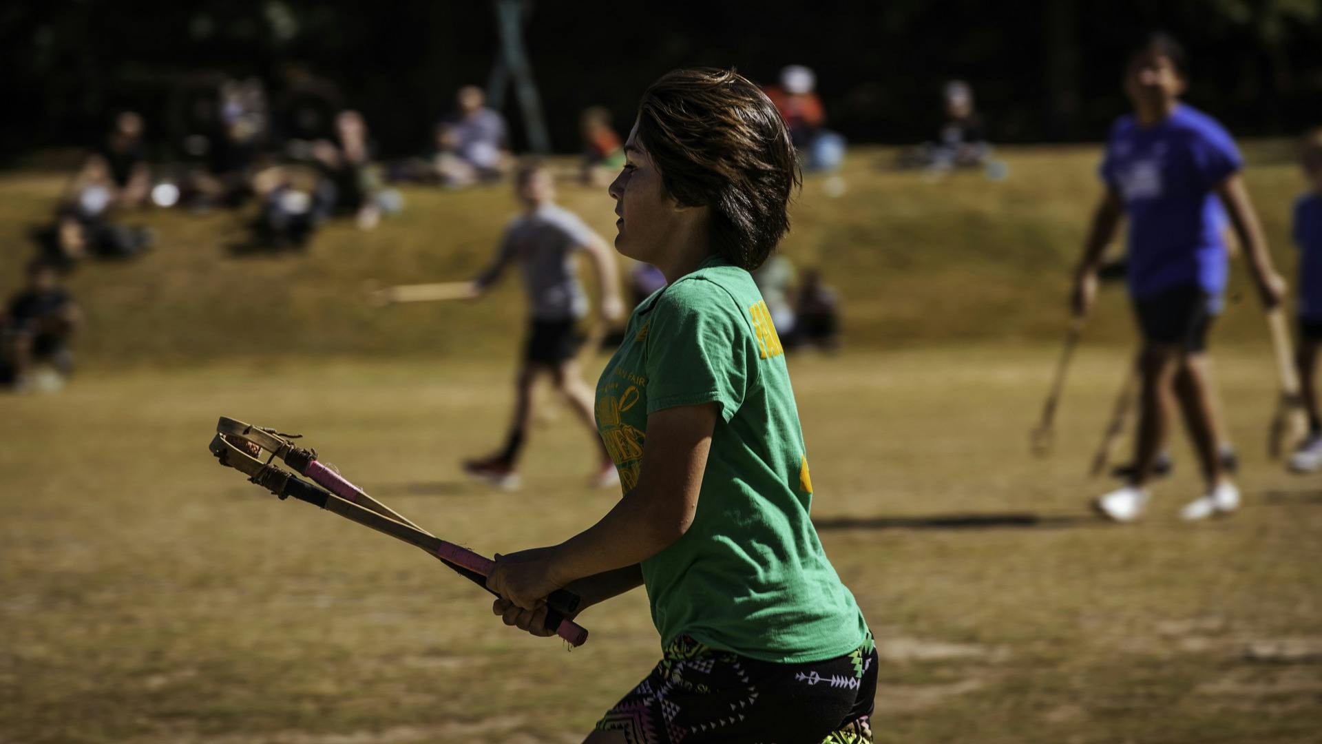 A Stickball player holds their sticks in front of them, while others run on the field in the background.
