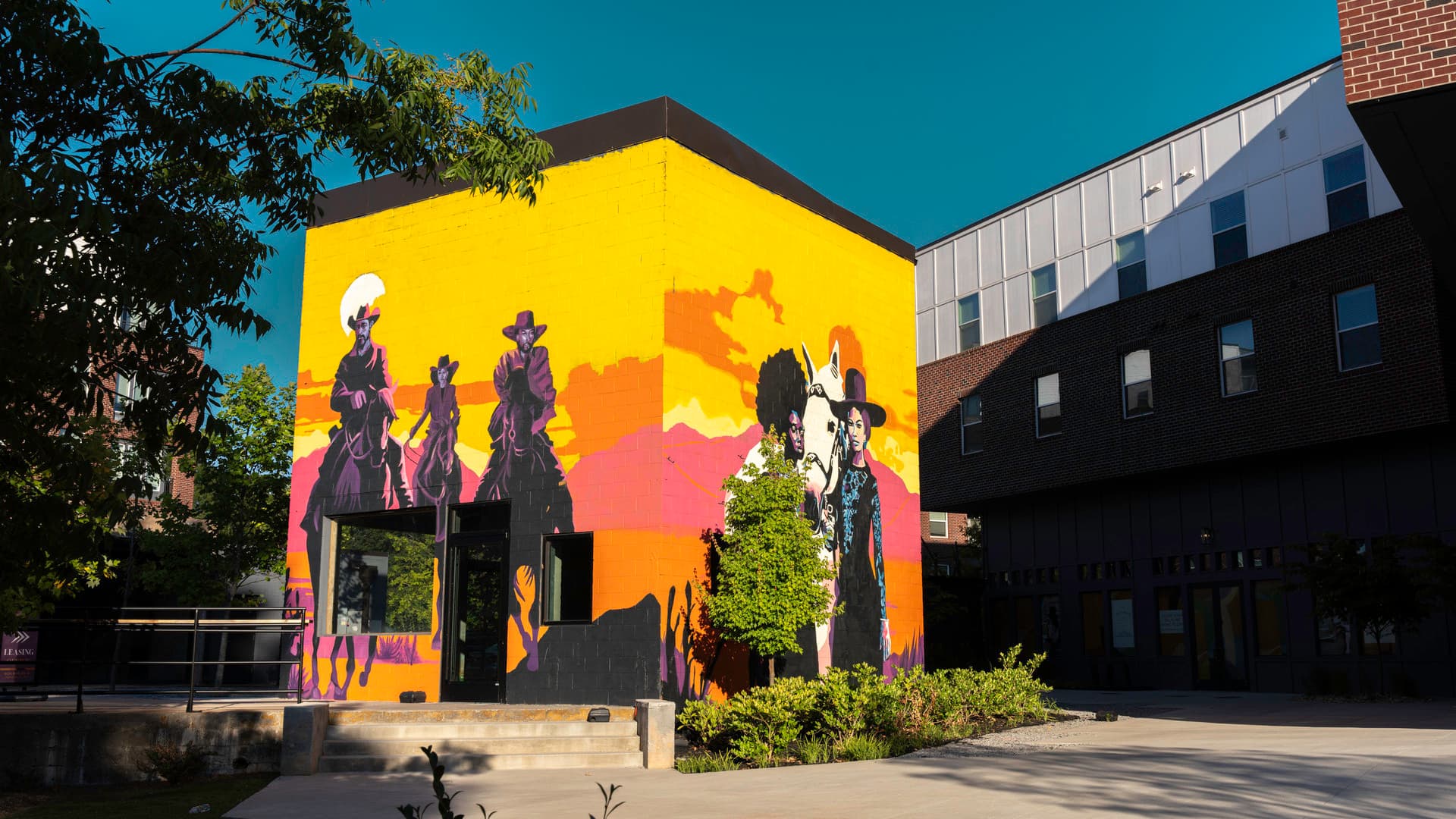 An image of two vibrantly colored murals on adjacent sides of a small building, surrounded by greenery and buildings. One mural features three figures on horseback wearing cowboy hats, the other showing two women standing with a horse, both set against a backdrop of mountains and sky.