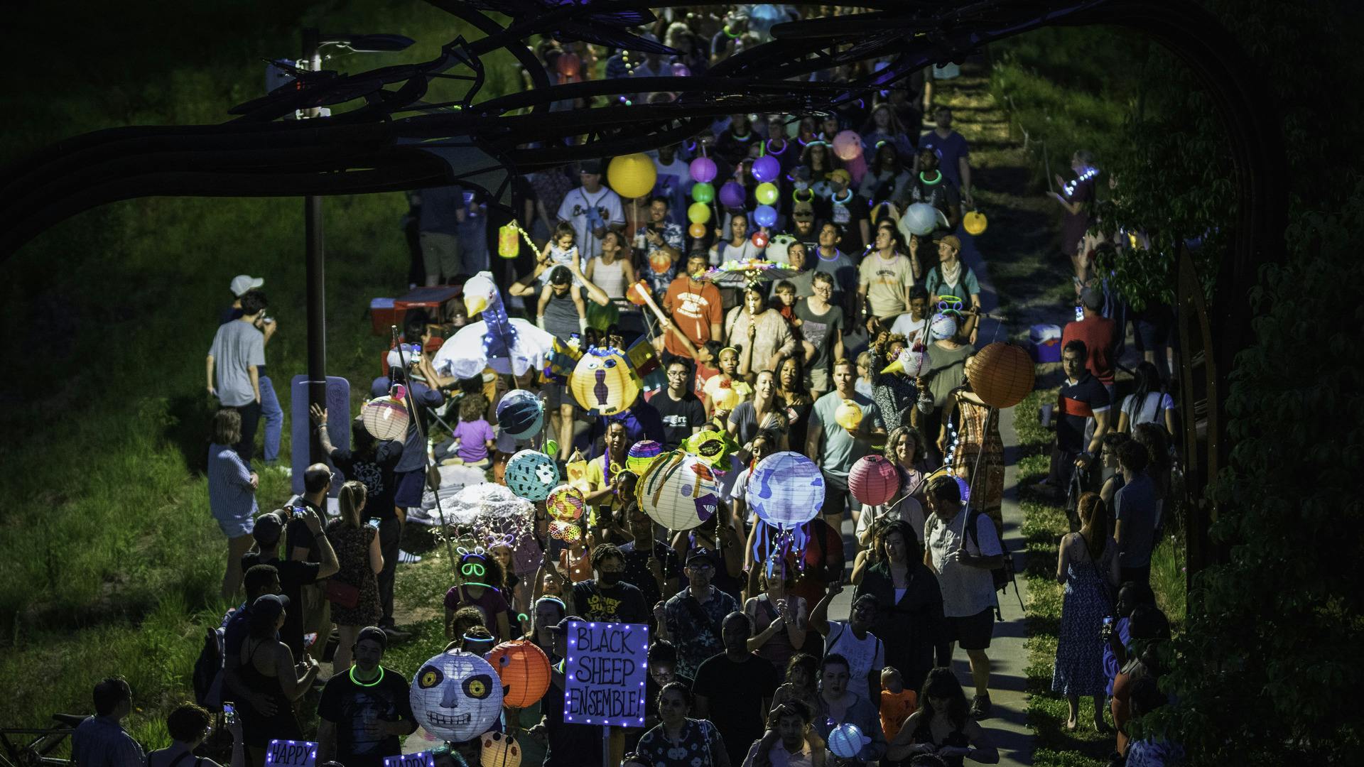 A crowd of people walk down the Beltline holding colorful, illuminated lanterns while a crowd watches on.