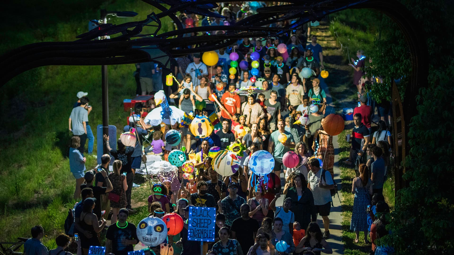 A crowd of people walk down the Beltline holding colorful, illuminated lanterns while a crowd watches on.