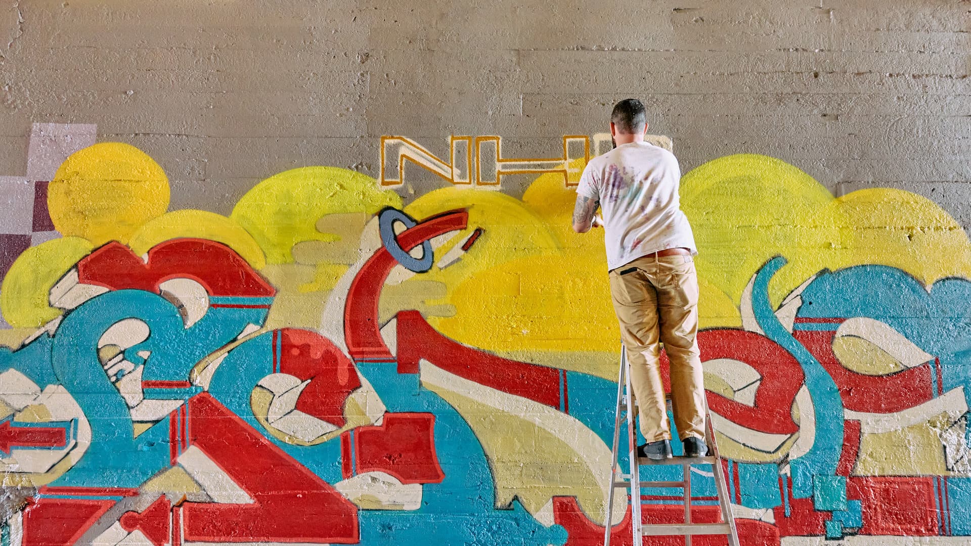 A man stands on a ladder with his back to the camera while he works on a graffiti mural.