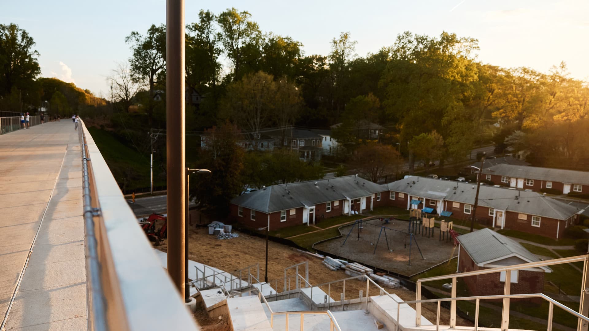 Elevated walkway overlooking residential neighborhood with red brick houses and a playground, surrounded by trees at sunset.