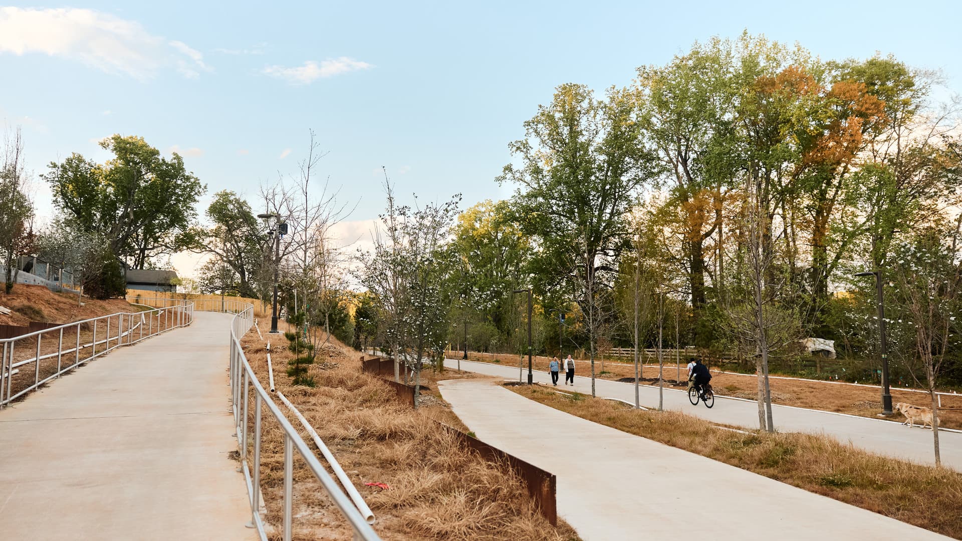Paved pathways with railings, surrounded by trees. A cyclist, walkers, and a dog are visible on the right path.