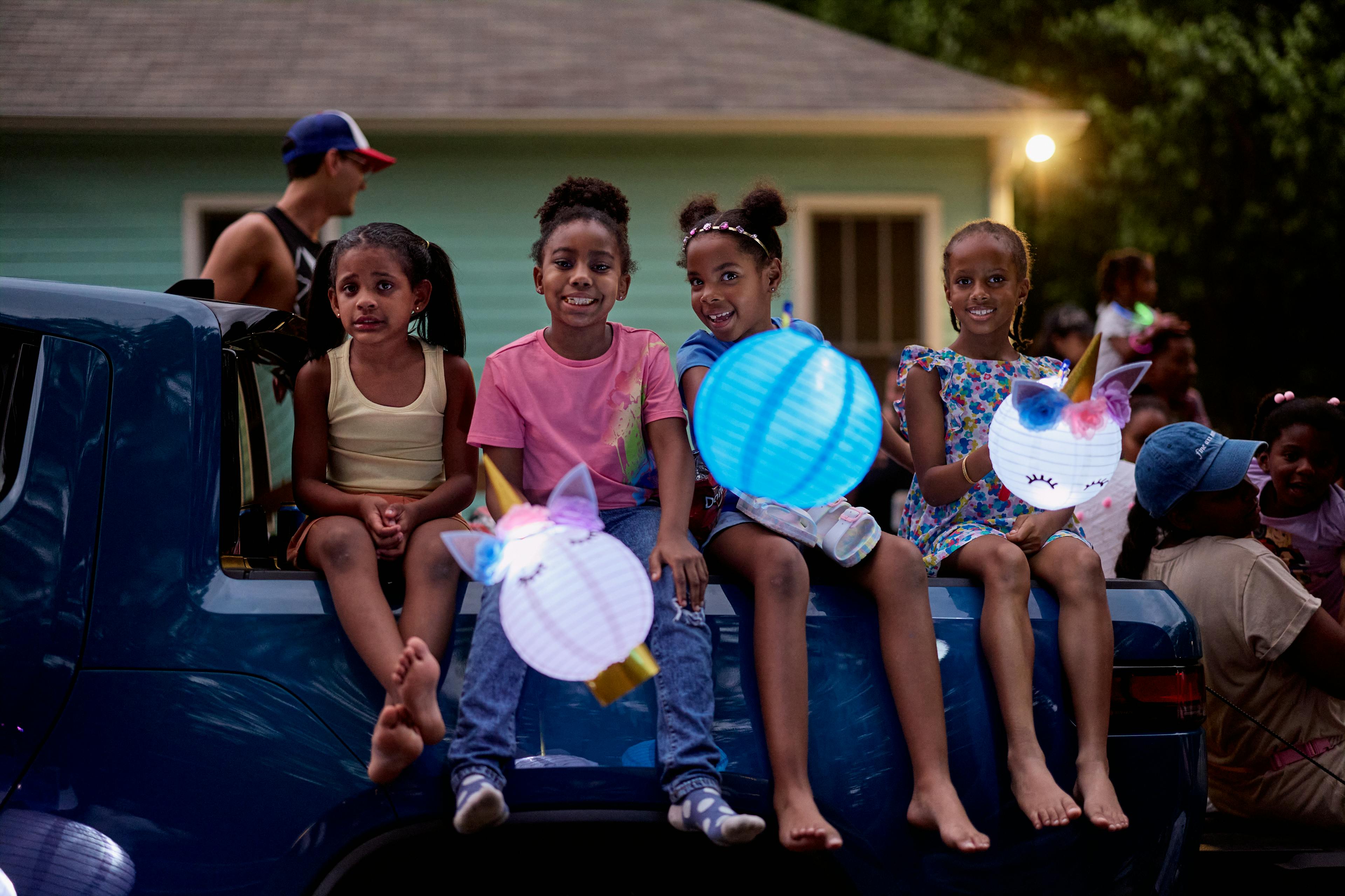 A group of young girls sit on the back of a truck holding homemade lanterns and smiling.