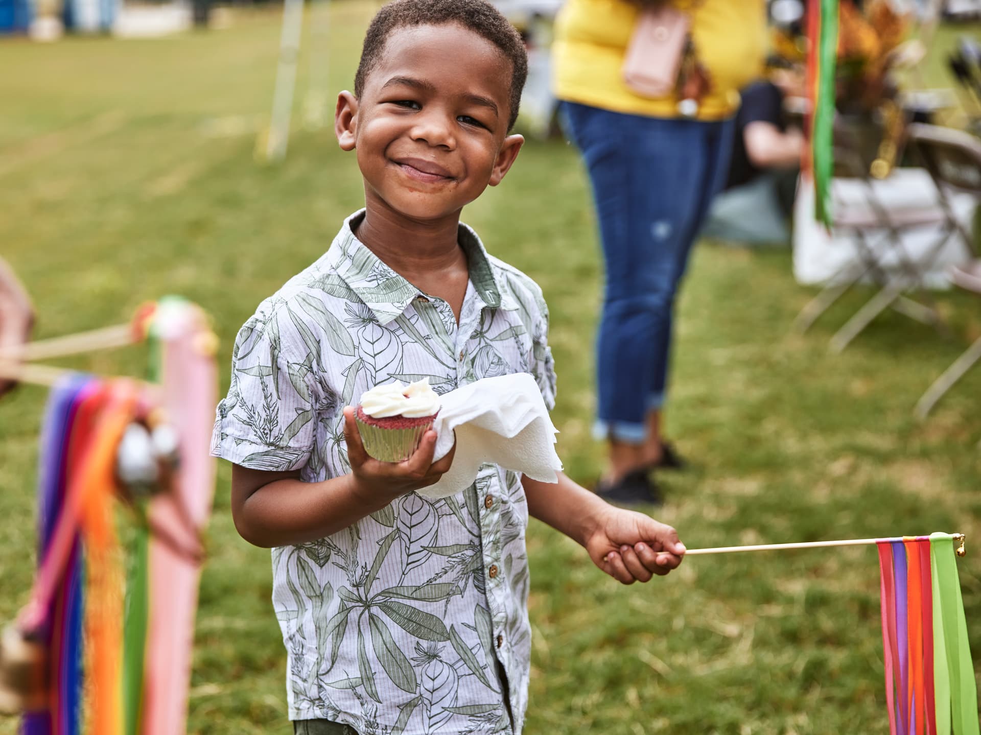 A smiling boy holds a cupcake and colorful ribbon stick outdoors, wearing a patterned shirt on a grassy field with people in the background.
