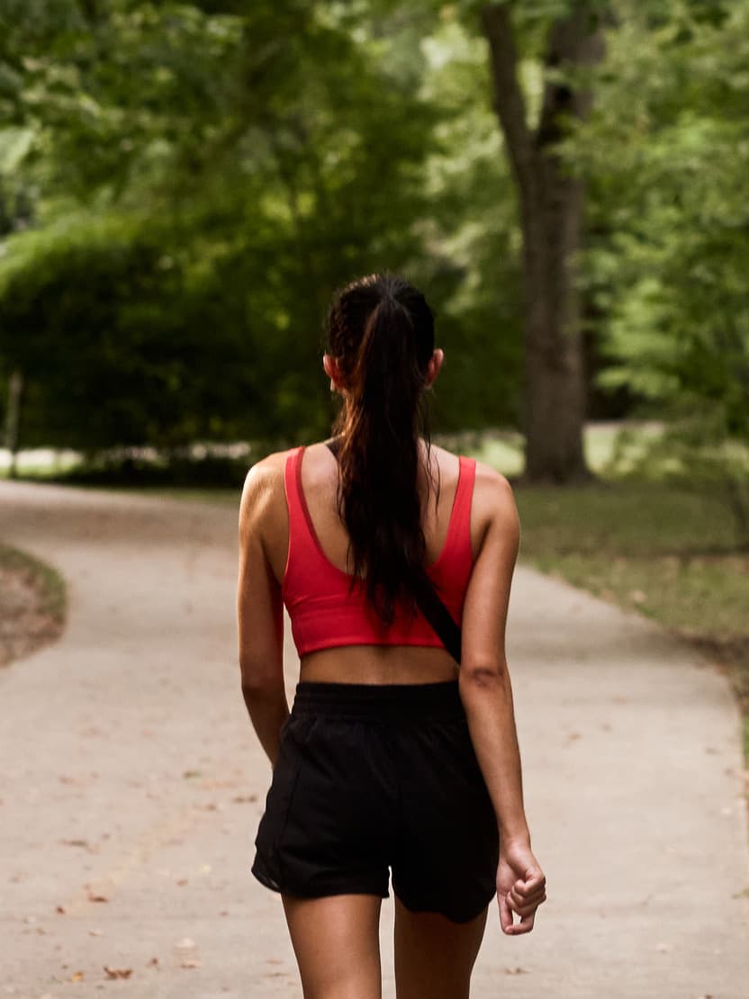 A woman in a red sports top and black shorts walks on a paved path tucked among lush greenery.