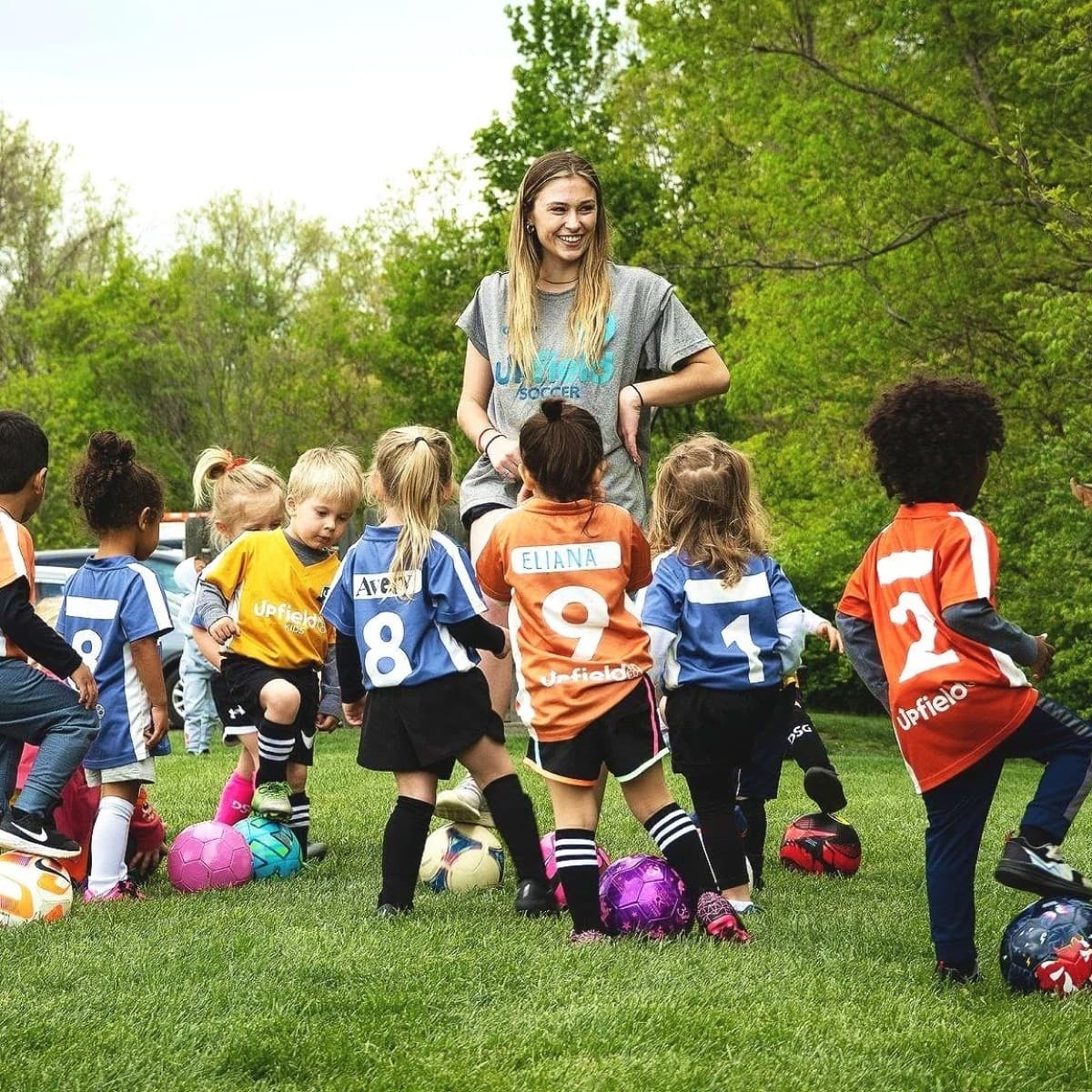 Young children in colorful sports jerseys gather around a coach on a grassy field, each with a soccer ball, ready to play.