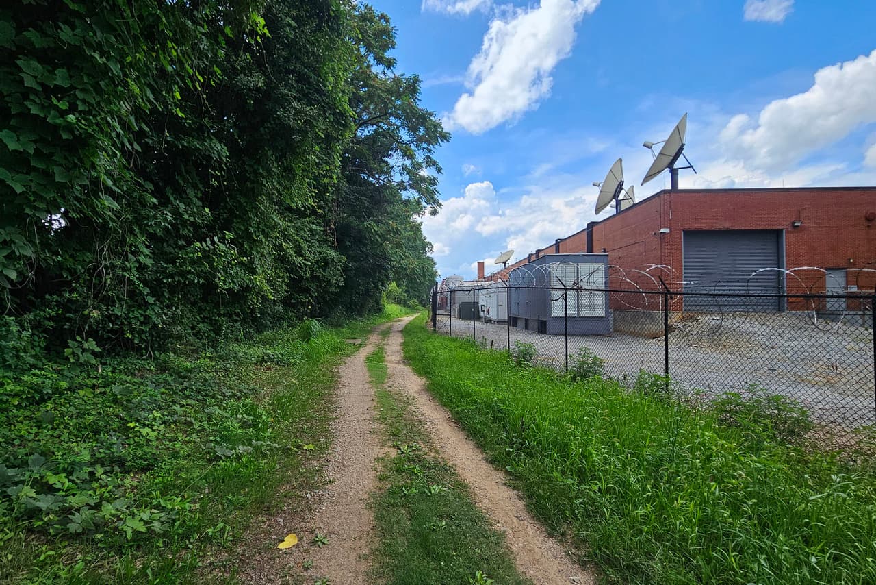 A worn dirt path runs between a line of thick trees and an industrial building.